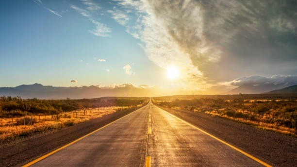 Estrada reta em meio a uma paisagem árida, com o sol baixo no horizonte e montanhas ao fundo